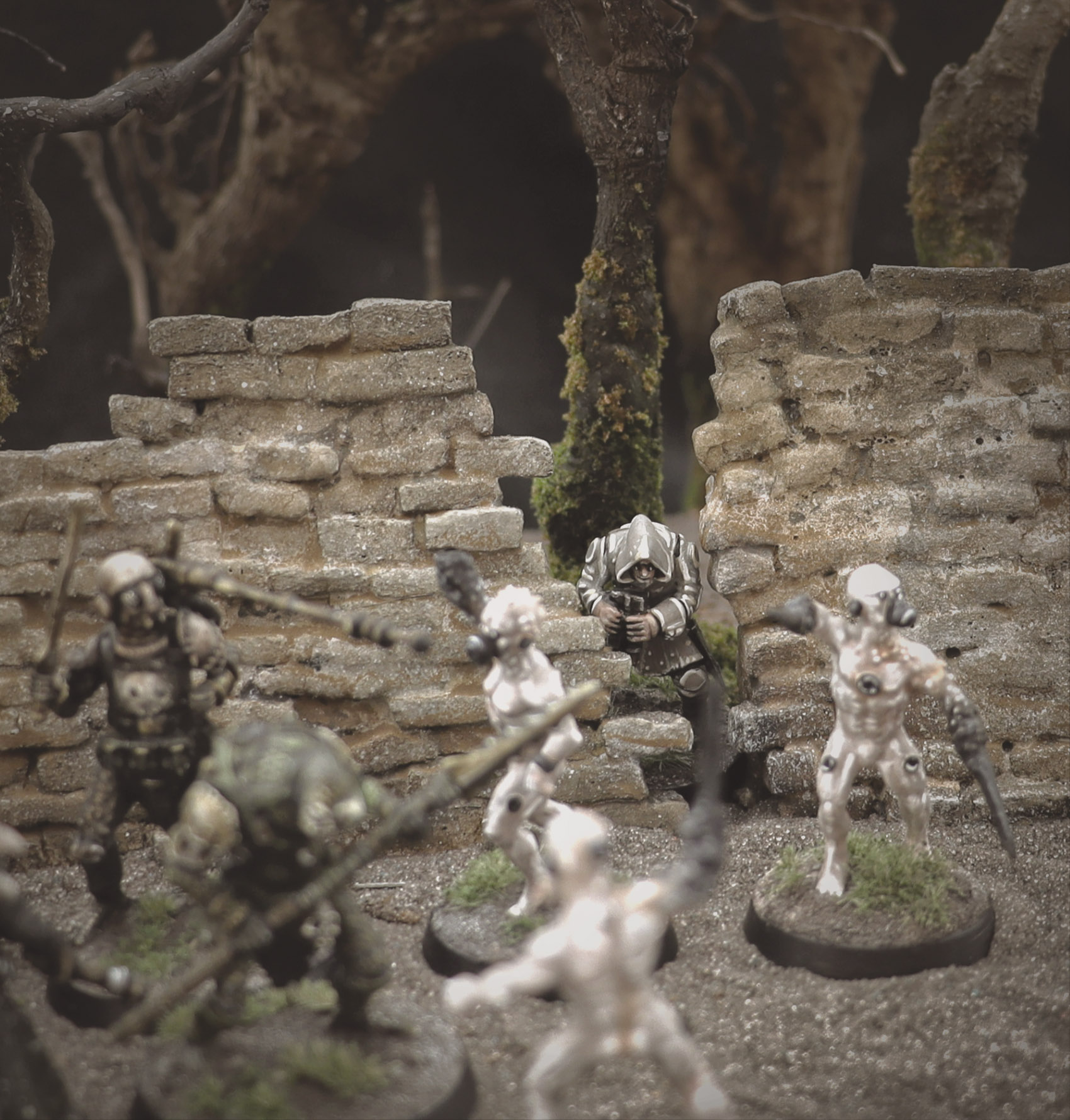 A photo of fully painted miniature figures and terrain: a band of hunters is fighting a group of white-skinned, biomechanical humanoids, while another figure crouches watching from behind a ruined wall